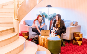 Two women sit in a lounge chair setup at base of curved staircase